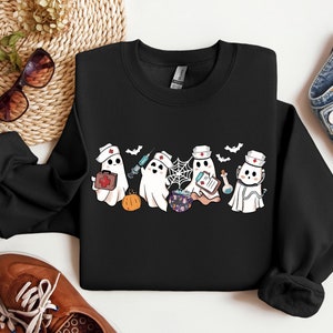 May include: Black sweatshirt with a graphic of four cute ghost nurses wearing red crosses and holding medical supplies. The ghosts are wearing white hats and have a friendly expression. The graphic is surrounded by bats and a spider web.