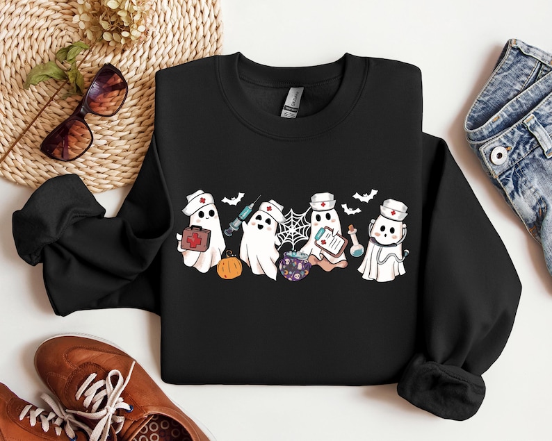 May include: Black sweatshirt with a graphic of four cute ghost nurses wearing red crosses and holding medical supplies. The ghosts are wearing white hats and have a friendly expression. The graphic is surrounded by bats and a spider web.
