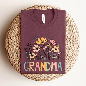 Puede incluir: Una camiseta color granate con un dise&ntilde;o floral y la palabra "GRANDMA" en letras de colores.