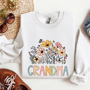 Puede incluir: Sudadera blanca con un dise&ntilde;o floral y la palabra "Abuela" en letras de colores.