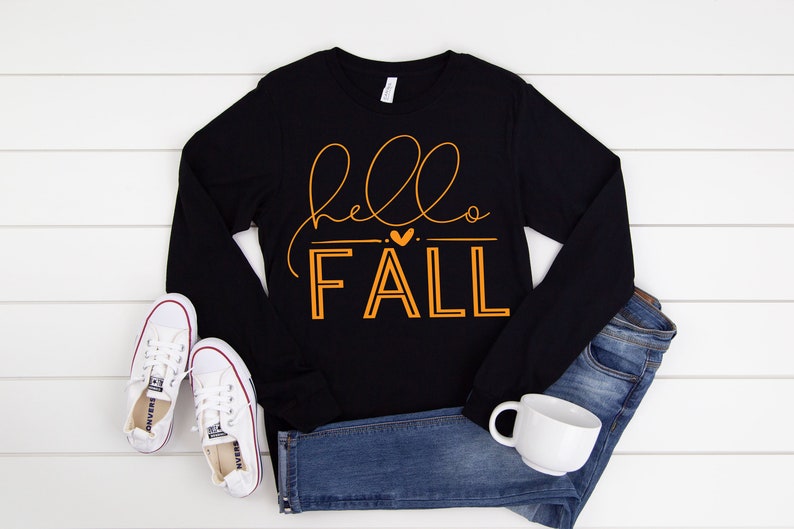 May include: Black long-sleeve shirt with the text "hello fall" in orange lettering with a small heart between the words. The shirt is laid flat on a white wood surface with a pair of white sneakers and blue jeans.