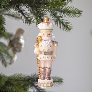 Op de afbeelding: Een roze en gouden glazen kerstbal met een notenkraker met een witte baard en een gouden staf. De kerstbal hangt aan een kerstboomtak.