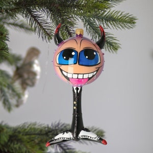 Puede incluir: Un adorno de Navidad de vidrio con forma de personaje de dibujos animados con una cara rosa, ojos azules, una amplia sonrisa y una lengua rosa que sobresale. El personaje lleva un traje negro y zapatos negros con detalles blancos. El adorno tiene cuernos rojos y está colgado de un árbol de Navidad.