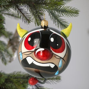 Puede incluir: Un adorno navideño negro y blanco con forma de diablo con nariz roja, ojos rojos y lágrimas azules en forma de gota. El adorno tiene cuernos amarillos y una cola roja con purpurina.