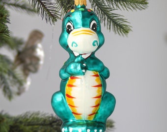 May include: A teal blue glass Christmas ornament shaped like a dinosaur with yellow and white stripes and a white face with black eyes.