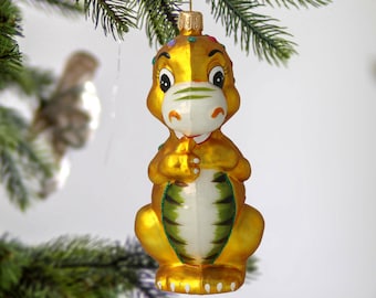 May include: A gold glass Christmas ornament shaped like a dinosaur with green and white stripes. The ornament has a white face with black eyes and a red mouth.