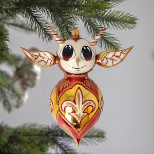 Puede incluir: Un adorno de Navidad de vidrio caprichoso con forma de criatura con ojos grandes, acentos rojos y dorados y un diseño de flor de lis.