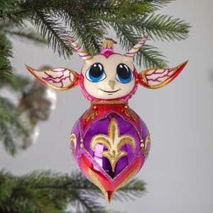Puede incluir: Un adorno navideño caprichoso con forma de criatura de dibujos animados con grandes ojos azules, cuernos y alas. El adorno presenta una combinación de colores rojo, morado y dorado con detalles de purpurina. Cuelga de una rama verde de árbol de Navidad.