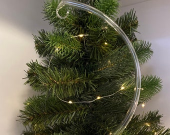 Op de afbeelding: Een heldere glazen displaystandaard voor kerstboomornamenten. De standaard heeft een gebogen arm met een haak aan de bovenkant en een cirkelvormige basis. De standaard staat voor een kleine kunstkerstboom met lichtjes.
