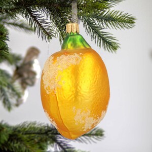 Puede incluir: Un adorno de cristal con forma de lim&oacute;n, cuerpo amarillo vibrante y parte superior verde. La superficie presenta un acabado texturizado con purpurina blanca escarchada en los laterales. Incluye una tapa de metal dorado con una peque&ntilde;a anilla.