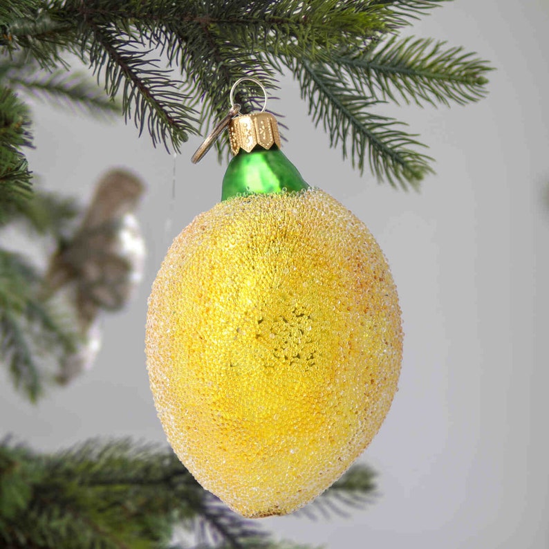Op de afbeelding: Een gele glazen kerstbal in de vorm van een citroen met een groene bovenkant. De bal is bedekt met kleine, glinsterende kralen.