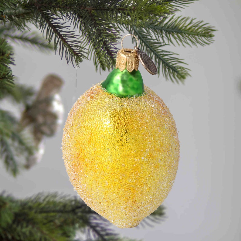 Op de afbeelding: Een gele glazen citroenversiering met een groene bovenkant en een gouden ornamentbevestiging. De versiering is bedekt met kleine, witte, glinsterende stippen.
