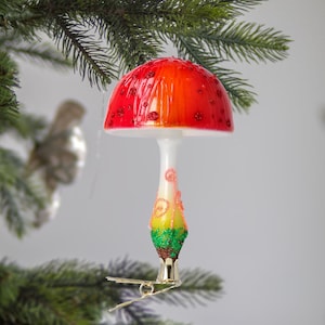May include: A red and white glass mushroom Christmas ornament with green and brown details. The ornament has a glitter finish and is designed to hang on a Christmas tree.