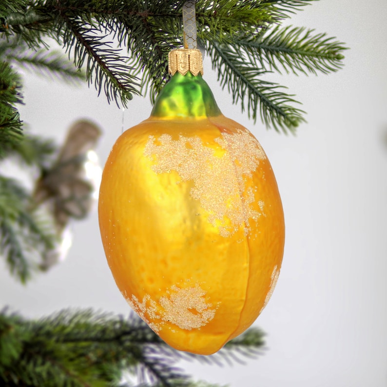 Puede incluir: Un adorno de &aacute;rbol de Navidad de vidrio con forma de lim&oacute;n, acabado en amarillo vibrante y detalles de purpurina dorada texturizada. El adorno cuenta con una tapa verde en la parte superior con un gancho plateado, colgado de una rama de pino.