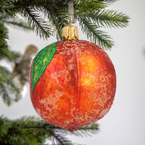 Puede incluir: Un adorno navideño de vidrio rojo con forma de melocotón con una hoja verde brillante. El adorno tiene una superficie texturizada que parece estar cubierta de escarcha.