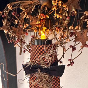 May include: A decorative candle holder wrapped in orange and black checkered fabric, featuring a black ribbon bow and natural fiber accents. A small, glowing yellow LED candle sits on top, surrounded by a rustic wreath of brown berries and leaves.