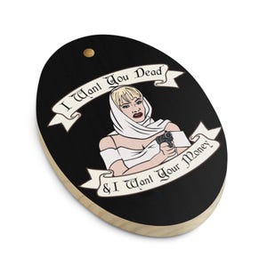 May include: Oval wooden plaque with a black background featuring a woman holding a gun. The text "I Want You Dead & I Want Your Money" is displayed on banners above and below the woman.