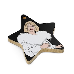 May include: Star-shaped wooden ornament featuring a pop art style illustration of a woman with blonde hair and a white fur collar. The background is black, and the ornament has a natural wood edge with a small gold-colored rivet at the top.
