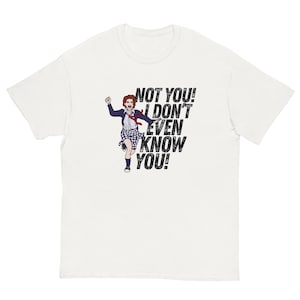 以下が含まれることがあります： 白いTシャツに、走っている女性の漫画の画像と「NOT YOU! I DON'T EVEN KNOW YOU!」というテキストが書かれています