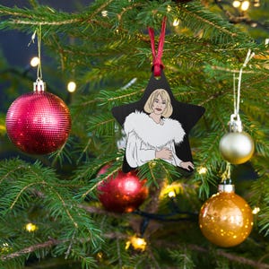 Op de afbeelding: Een zwarte, stervormige kerstversiering met een afbeelding van een vrouw in een witte jas met bontkraag. De versiering hangt aan een rood lint in een kerstboom, omringd door rode en gouden ballen.