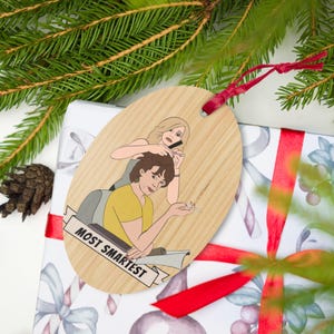 May include: Wooden Christmas tree decoration with a cartoon illustration of two people. One is combing the other's hair while holding a phone. The other is holding a cigarette. The text "MOST SMARTEST" is on a banner.
