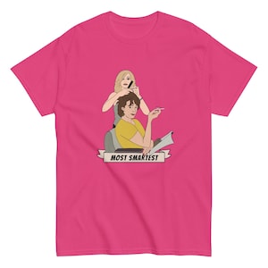 May include: A vibrant pink t-shirt with a cartoon image of two people, one cutting the other's hair. The text "MOST SMARTEST" is displayed on a banner. The t-shirt is made of a soft material and is suitable for casual wear.