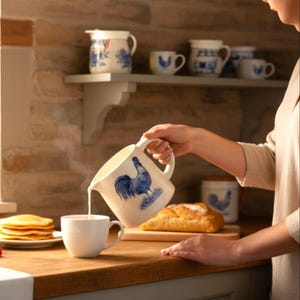 May include: A white ceramic jug with a blue rooster design pours liquid into a white mug. Matching mugs and a jug are on a shelf. Pancakes and a loaf of bread are on the wooden countertop.
