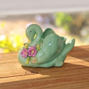 May include: A light green ceramic swan planter featuring a sculpted cluster of pink and yellow flowers on its chest. The swan has a smooth, glossy finish, a curved neck, and a hollow opening on its back for holding small items or plants.