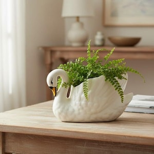 Op de afbeelding: Een witte keramische zwaan planter gevuld met groene planten. De zwaan heeft een gebogen nek, een gele snavel en zwarte ogen. De planter staat op een houten tafel.