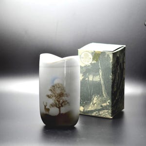 May include: A white ceramic vase with a curved top edge, featuring a brown silhouette of a deer standing under a tree with a pale blue moon above. Beside it stands a rectangular cardboard box with a detailed black and white landscape print.