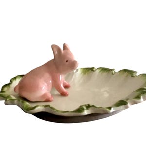 May include: A ceramic dish shaped like a leaf, with a light pink pig figurine perched on top. The leaf-shaped dish has a white interior and a green, ruffled edge. The pig is in a sitting position.