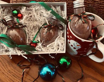 May include: Two clear glass ornaments filled with brown powder, packaged in a white box with green and red ribbon. The ornaments are shaped like light bulbs and are sitting on white shredded paper. A third ornament is in a red and white mug with a snowman design.