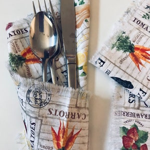 May include: A set of silverware wrapped in a white fabric napkin with a farmhouse-style print featuring vegetables and the text "ORGANIC", "LOCALLY GROWN", "CARROTS", "TOMATOES", "FRESH LOCAL", "STRAWBERRIES", "FARMERS MARKET", and "FA RS".