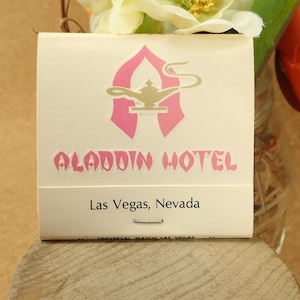 May include: A vintage matchbook cover for the Aladdin Hotel in Las Vegas, Nevada. The cover features a pink "A" with a gold genie lamp inside. The text "Aladdin Hotel" is in pink letters and "Las Vegas, Nevada" is in black letters.