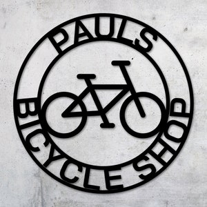 May include: Black and white circular sign with the text "Paul's Bicycle Shop" around the edge. A bicycle silhouette is in the center of the sign.