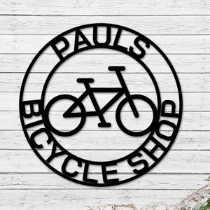 May include: Black and white metal sign with the text "Paul's Bicycle Shop" in a circle around a silhouette of a bicycle.