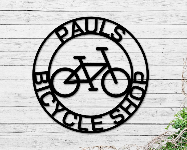 May include: Black and white metal sign with the text "Paul's Bicycle Shop" in a circle around a silhouette of a bicycle.