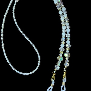 Könnte beinhalten: Eine Perlen-Brillenkette mit filigranem Design. Die Kette besteht aus kleinen, weißen Perlen und größeren, irisierenden Kristallperlen. Goldfarbene Verschlüsse befestigen die Kette an der Brille. Der Hintergrund ist schwarz.