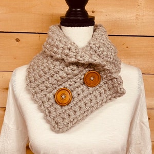 May include: A light gray, chunky-knit cowl neck warmer with two large, round, wooden buttons. The neck warmer is displayed on a white mannequin, showcasing its textured, handmade appearance. The background is a light brown wooden wall.