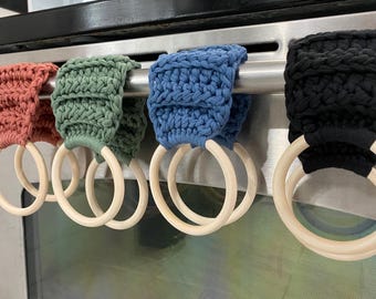 May include: Four handmade crochet kitchen towel holders in coral, green, blue, and black. Each holder has a wooden ring for hanging. The holders are attached to a stainless steel bar, likely on an oven.
