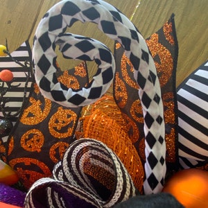 Puede incluir: Una corona de Halloween con cinta a cuadros blanco y negro, cinta de brillo naranja y negro y cinta de malla naranja. La corona tambi&eacute;n tiene cinta a rayas blanco y negro, cinta con estampado de calabaza negro y naranja y un remolino a cuadros blanco y negro.