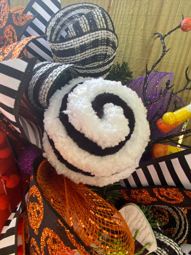 Puede incluir: Una corona de Halloween con cinta a rayas blancas y negras, un remolino blanco y negro, malla naranja y negra y malla morada y negra.