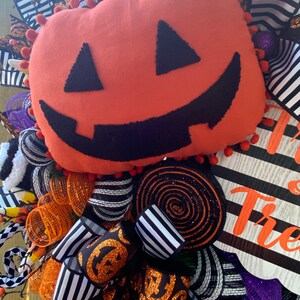 Puede incluir: Una corona de Halloween con una cinta a rayas blancas y negras, malla naranja y negra, y una gran calabaza de fieltro naranja con una cara sonriente negra. La corona tambi&eacute;n incluye un letrero que dice "Trick or Treat".