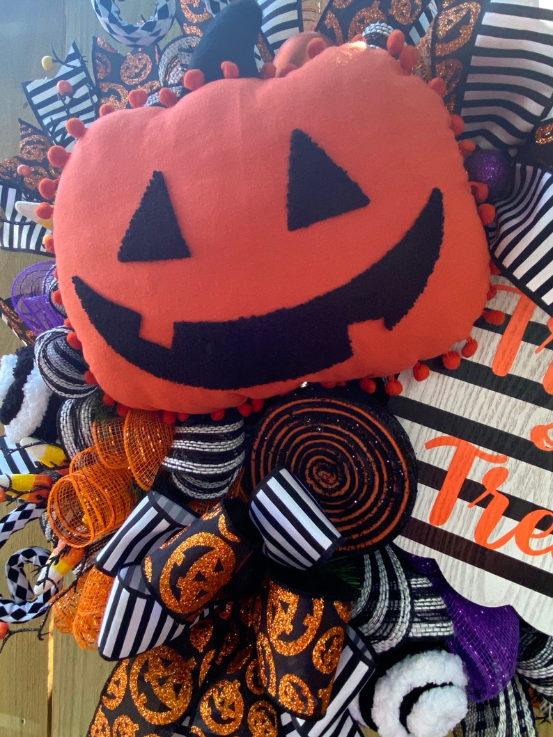 Puede incluir: Una corona de Halloween con una cinta a rayas blancas y negras, malla naranja y negra, y una gran calabaza de fieltro naranja con una cara sonriente negra. La corona tambi&eacute;n incluye un letrero que dice "Trick or Treat".