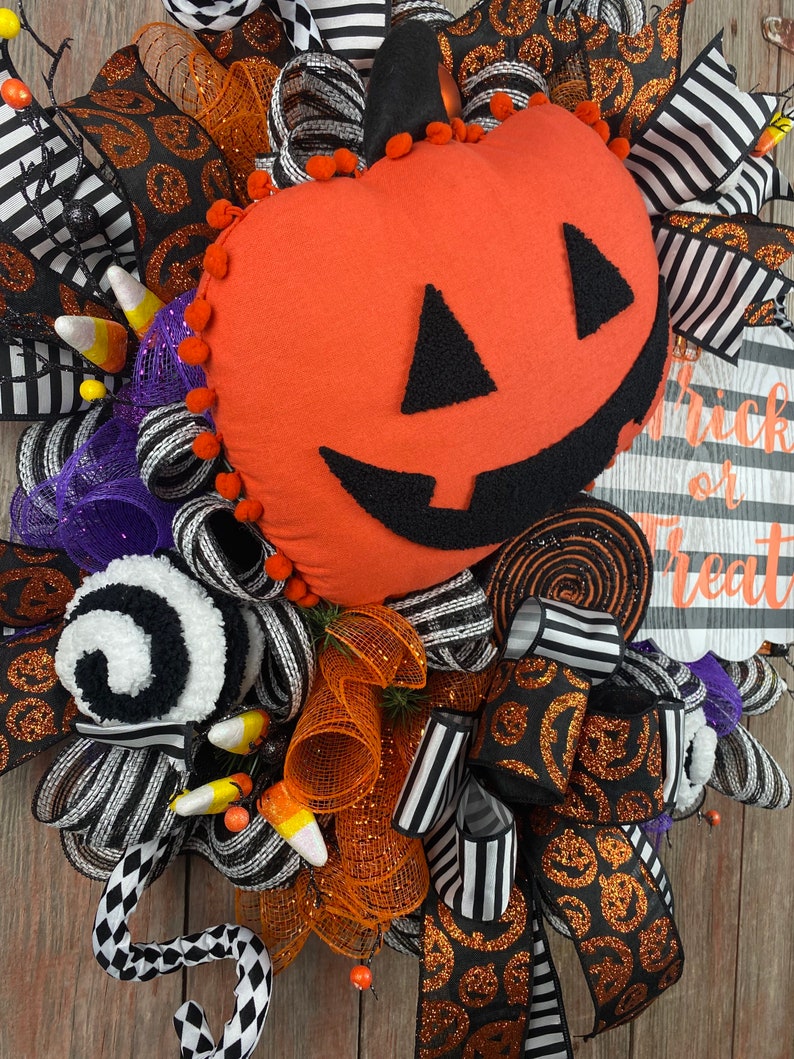 Puede incluir: Corona de Halloween con una cinta a rayas blancas y negras, una cinta de purpurina naranja y negra y una calabaza de fieltro con una cara de jack-o'-lantern. La corona tambi&eacute;n incluye ma&iacute;z dulce, decoraciones de espiral blancas y negras y un letrero que dice "Trick or Treat".