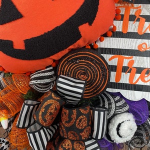 Puede incluir: Una corona de Halloween con fondo a rayas blanco y negro, un remolino negro y naranja, una cinta a rayas blanco y negro y una cinta negra con calabazas de purpurina naranja. La corona tambi&eacute;n presenta una gran almohada de calabaza naranja con una cara de calabaza negra y las palabras "Trick or Treat" en negro.