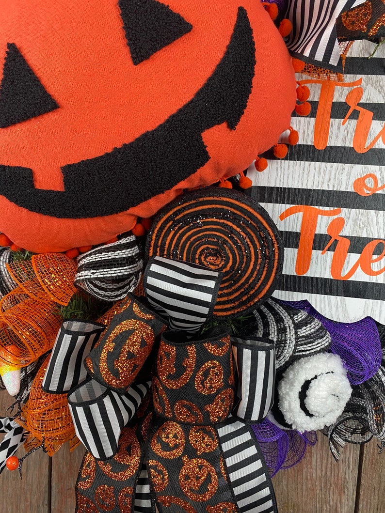 Puede incluir: Una corona de Halloween con fondo a rayas blanco y negro, un remolino negro y naranja, una cinta a rayas blanco y negro y una cinta negra con calabazas de purpurina naranja. La corona tambi&eacute;n presenta una gran almohada de calabaza naranja con una cara de calabaza negra y las palabras "Trick or Treat" en negro.