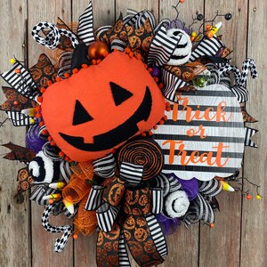 Puede incluir: Una corona de Halloween con una cinta a rayas blancas y negras, malla naranja y negra y una calabaza de felpa naranja con una cara negra. La corona tambi&eacute;n incluye un letrero que dice "Trick or Treat" en blanco y negro.