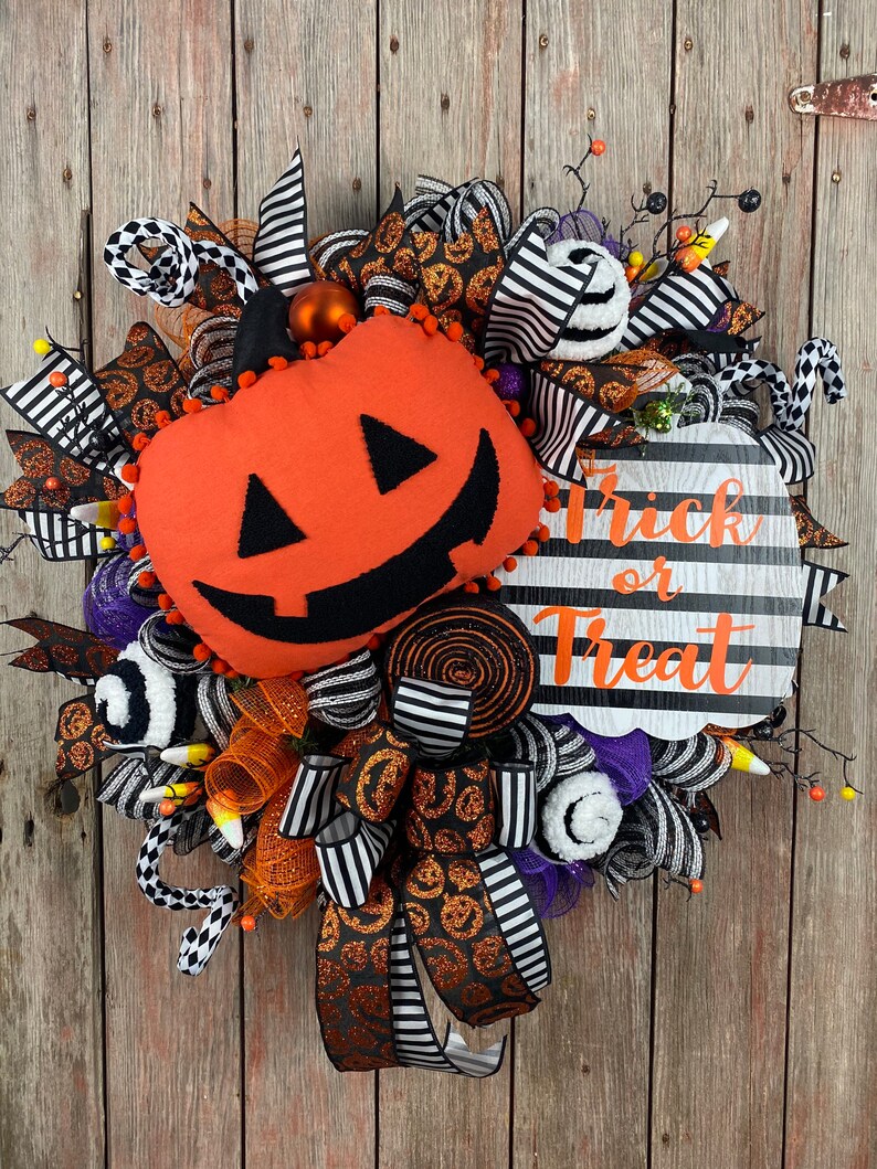 Puede incluir: Una corona de Halloween con una cinta a rayas blancas y negras, malla naranja y negra y una calabaza de felpa naranja con una cara negra. La corona tambi&eacute;n incluye un letrero que dice "Trick or Treat" en blanco y negro.