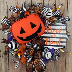 Puede incluir: Una corona de Halloween con una cinta a rayas blancas y negras, malla naranja y negra y una calabaza de peluche con una cara sonriente. La corona tambi&eacute;n incluye un letrero que dice "Trick or Treat".